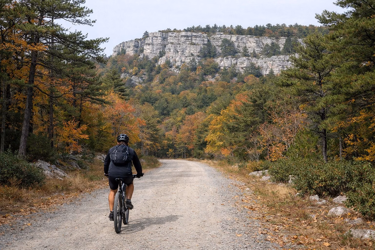 A cyclist in a helmet riding a gravel carriage road through autumn woods below cliffs