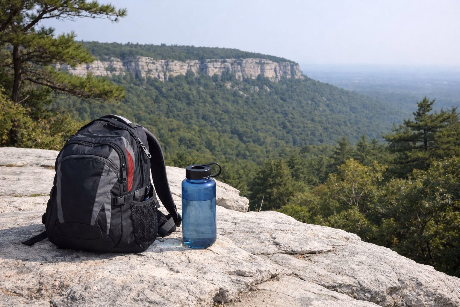 A daypack and water bottle on a pale rock ledge above a forested valley