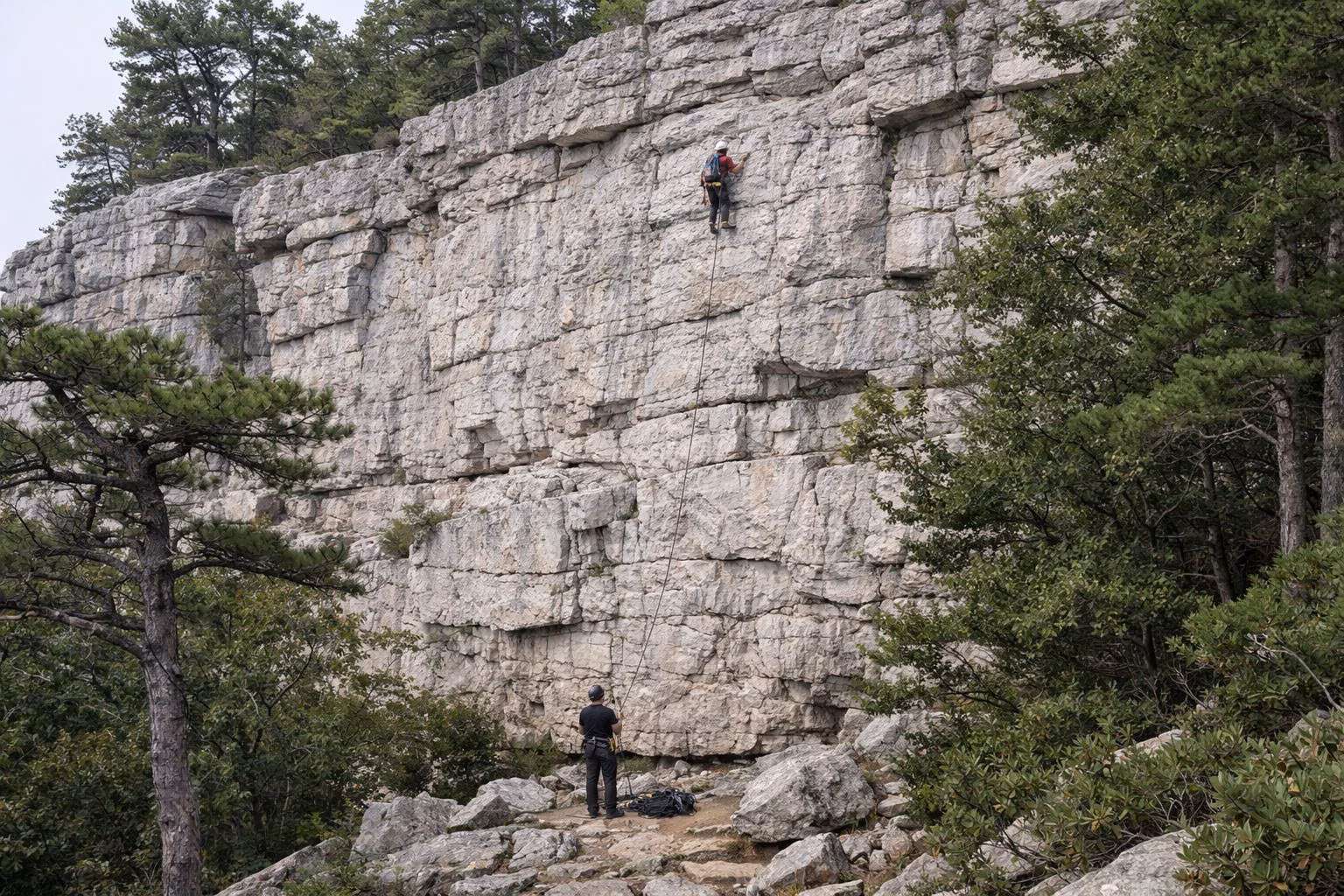 A roped climber high on a pale conglomerate cliff with a belayer at the base
